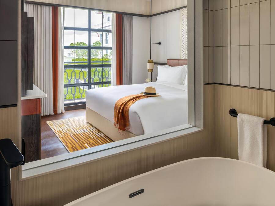 A hotel room with a large bed, a bathroom with a bathtub, and a view of greenery outside.