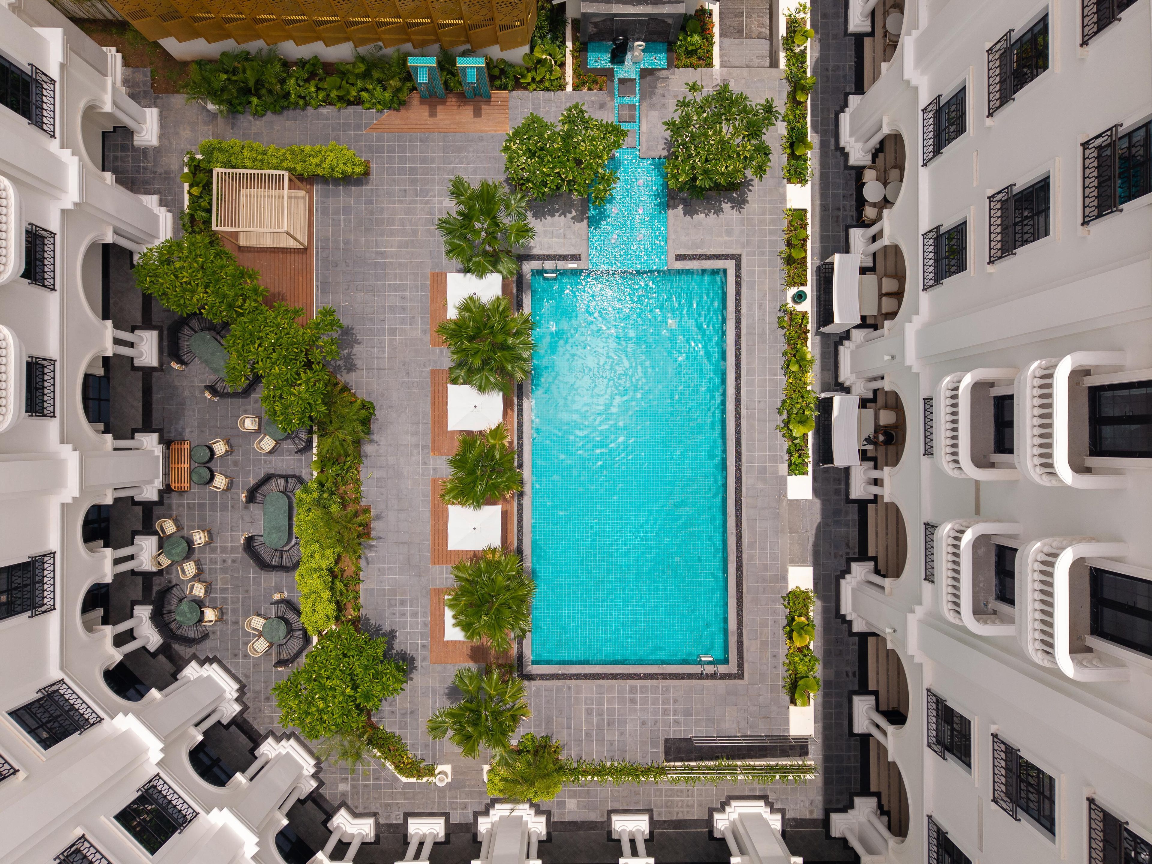 A rooftop pool area with a rectangular pool, surrounded by lounge chairs and tables, and bordered by greenery.