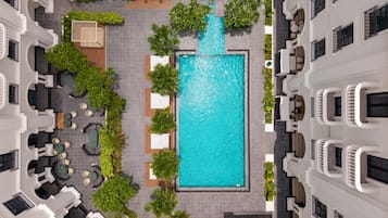 A rooftop pool area with a rectangular pool, surrounded by lounge chairs and tables, and bordered by greenery.