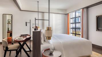 A modern hotel room with a large bed, a desk, a chair, a round side table, a mirror, and a large window with a view.