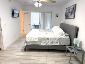Une chambre à coucher comprenant un lit, un téléviseur, un ventilateur de plafond, une table de chevet et une fenêtre avec des rideaux.