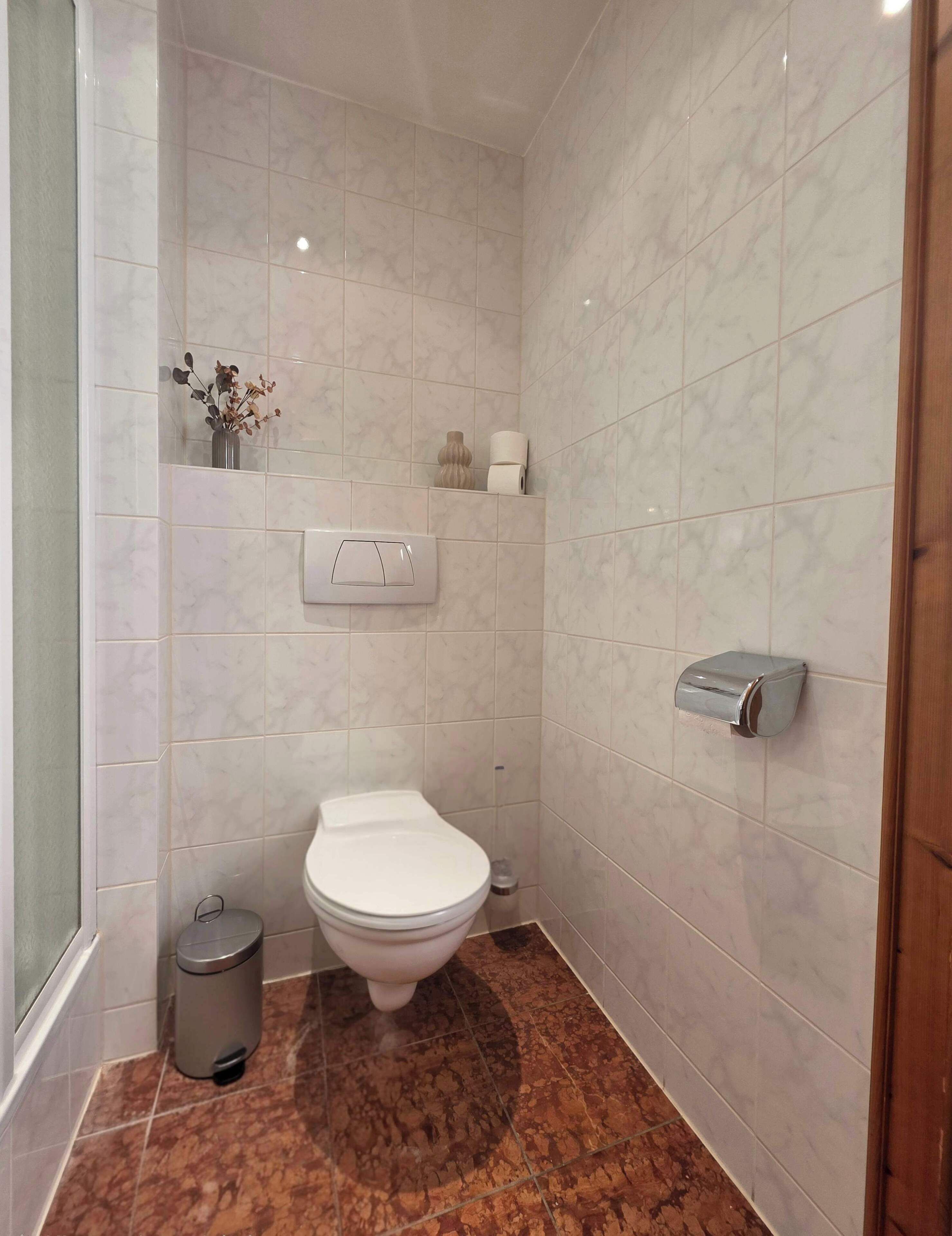 A bathroom with a toilet, a trash bin, a paper towel dispenser, and a small shelf with a vase and toilet paper.