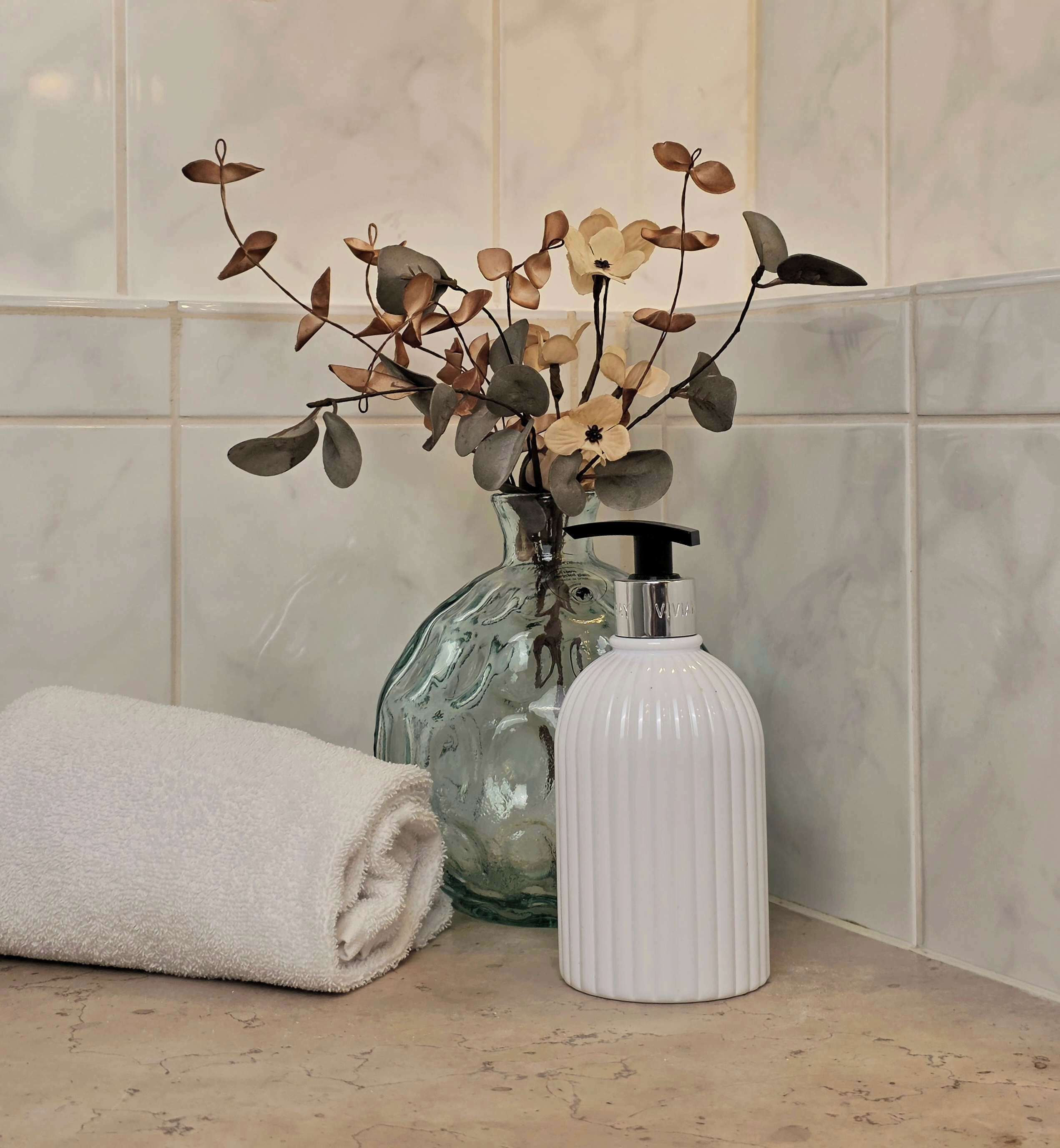 A bathroom with a towel, a glass vase with dried flowers, and a white bottle.