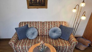 A living room with a patterned sofa, two blue cushions, a wooden coffee table, a floor lamp, and a wicker basket. - Alpine Retreat: Pet-Friendly Apartment w/ Free (Fischbachau)