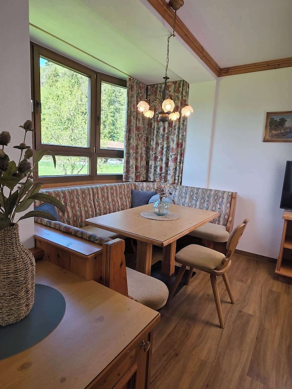 Une pièce chaleureuse avec une table à manger en bois, des bancs, un luminaire suspendu et une grande fenêtre avec des rideaux.