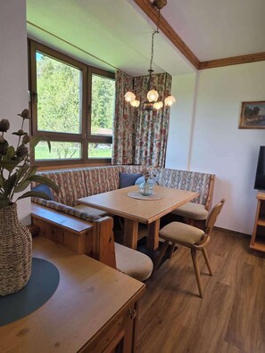 A cozy room with a wooden dining table, bench seating, a hanging light fixture, and a large window with curtains.