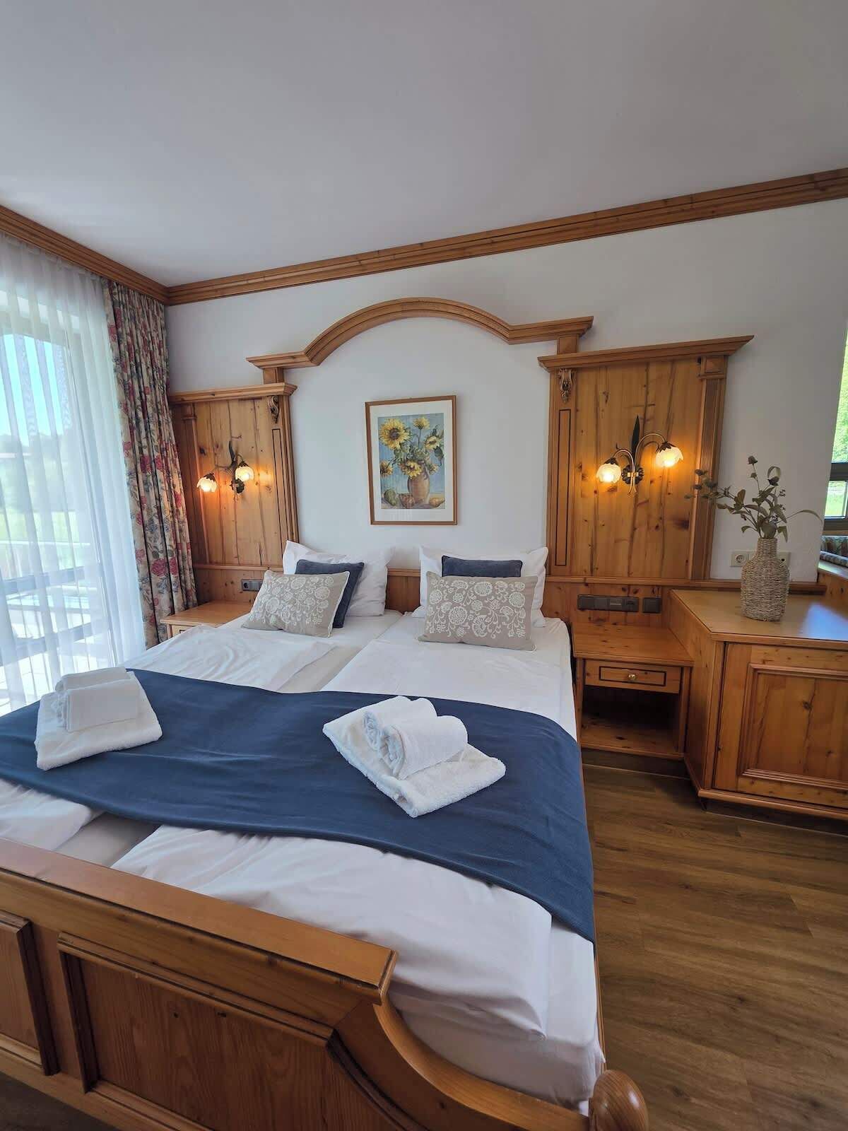 A hotel room with a large bed, wooden headboard, a painting above the bed, and a wooden nightstand with a vase.. Gemütliches Alpen-Apartment 4 in Wörnsmühl