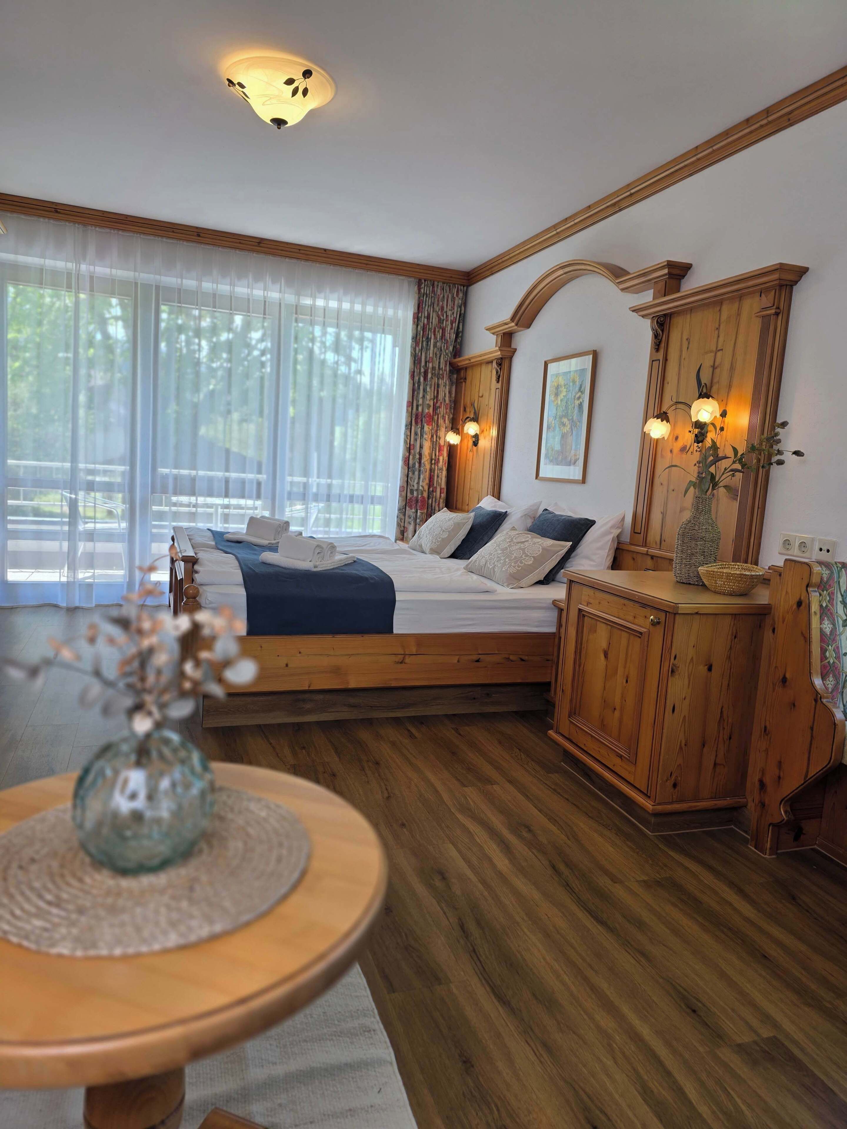 A bedroom with a wooden bed, a nightstand, a vase with flowers, and a window with sheer curtains.