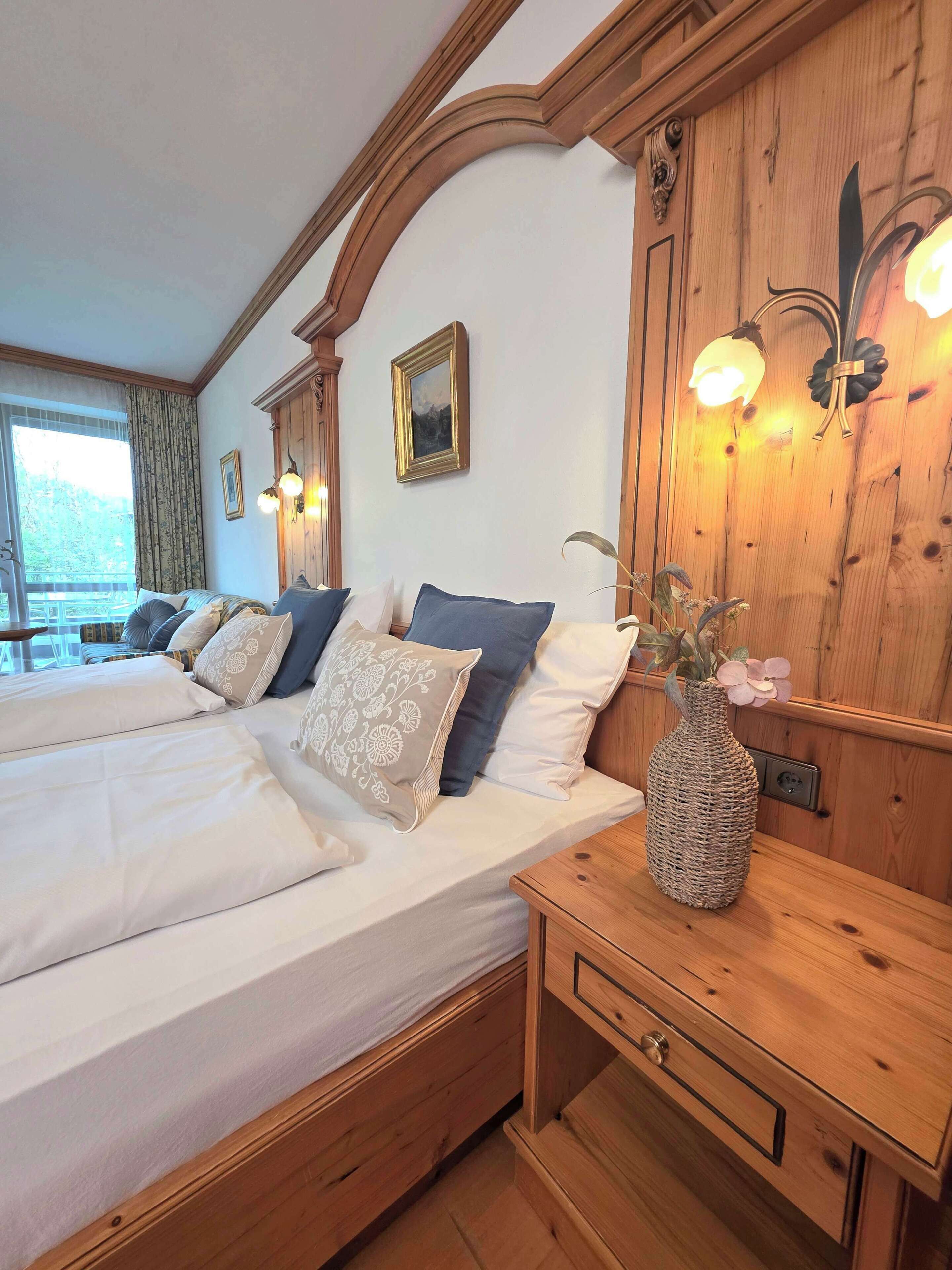 Een hotelkamer met een groot bed, een houten hoofdbord, een vaas met bloemen en een nachtkastje van hout met een lade.