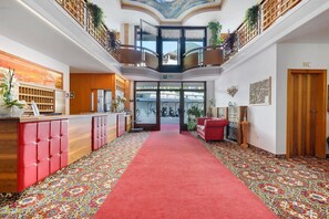Una hall d'albergo con tappeto rosso, una reception, un divano rosso e un balcone con piante.