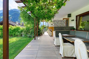 Un'area salotto esterna riparata, con tavoli e sedie, una pergola ricca di vegetazione e una vista sulle montagne sullo sfondo.