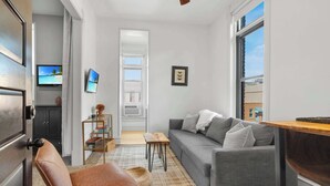A living room with a grey sofa, a brown leather chair, a small wooden table, a TV on the wall, and a window with a city view. - Downtown Palace Hotel Suite - Steps to Adventure (Salida)