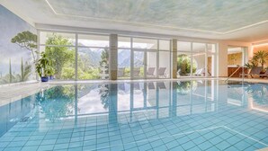 An indoor swimming pool with clear glass walls, a tiled floor, and a view of mountains through the windows. - Spacious Timmels Suite with South Balcony and Free Wellness & Pool Access (St. Leonhard in Passeier)
