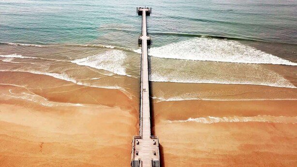Une jetée s’avançant dans l’eau, avec un bateau amarré à son extrémité, entourée d’une plage de sable et d’une mer calme.