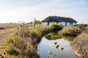 Une maison de campagne avec un toit de chaume, entourée d’un petit plan d’eau et d’une végétation luxuriante.