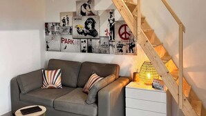 Una sala de estar con un sofá gris, una escalera de madera y una pared decorada con varios carteles.