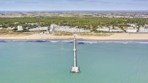 Vista aérea de una zona costera con un muelle largo que se adentra en el agua, rodeado de edificios y una playa.