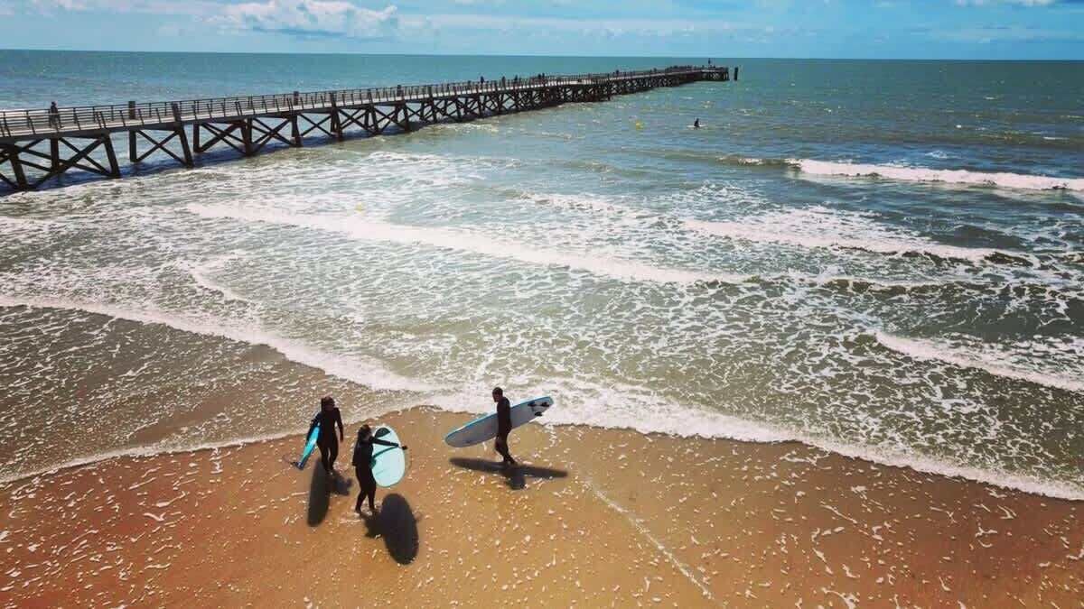 Une plage avec une jetée qui s’avance dans l’océan, des gens qui se promènent avec leurs planches de surf, et des vagues qui se rapprochent du rivage.