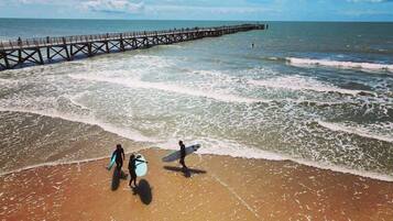 Une plage avec une jetée qui s’avance dans l’océan, des gens qui se promènent avec leurs planches de surf, et des vagues qui se rapprochent du rivage.