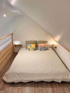 Een slaapkamer met een schuine plafond, houten bedframe, een bed met een patroon erover, twee kussens en een nachtkastje met een lamp.
