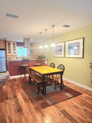 A dining area with a wooden table, chairs, and a rug. There is a kitchen in the background with cabinets and a refrigerator. - Palm Paradise 5 min to Downtown 5 min to the Beaches (St. Augustine)