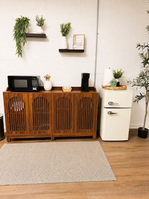 Une petite cuisine équipée d’un four à micro-ondes, d’une machine à café et d’un petit réfrigérateur. Des plantes ornent le mur, et un meuble en bois abrite un panier en osier.