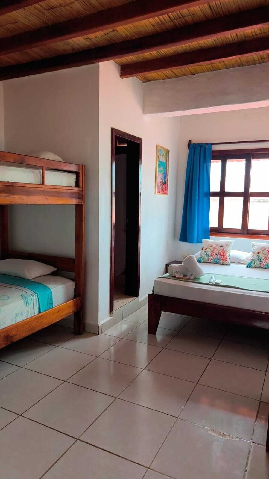 A room with a bunk bed, a single bed, a door, a window with blue curtains, and a painting on the wall.