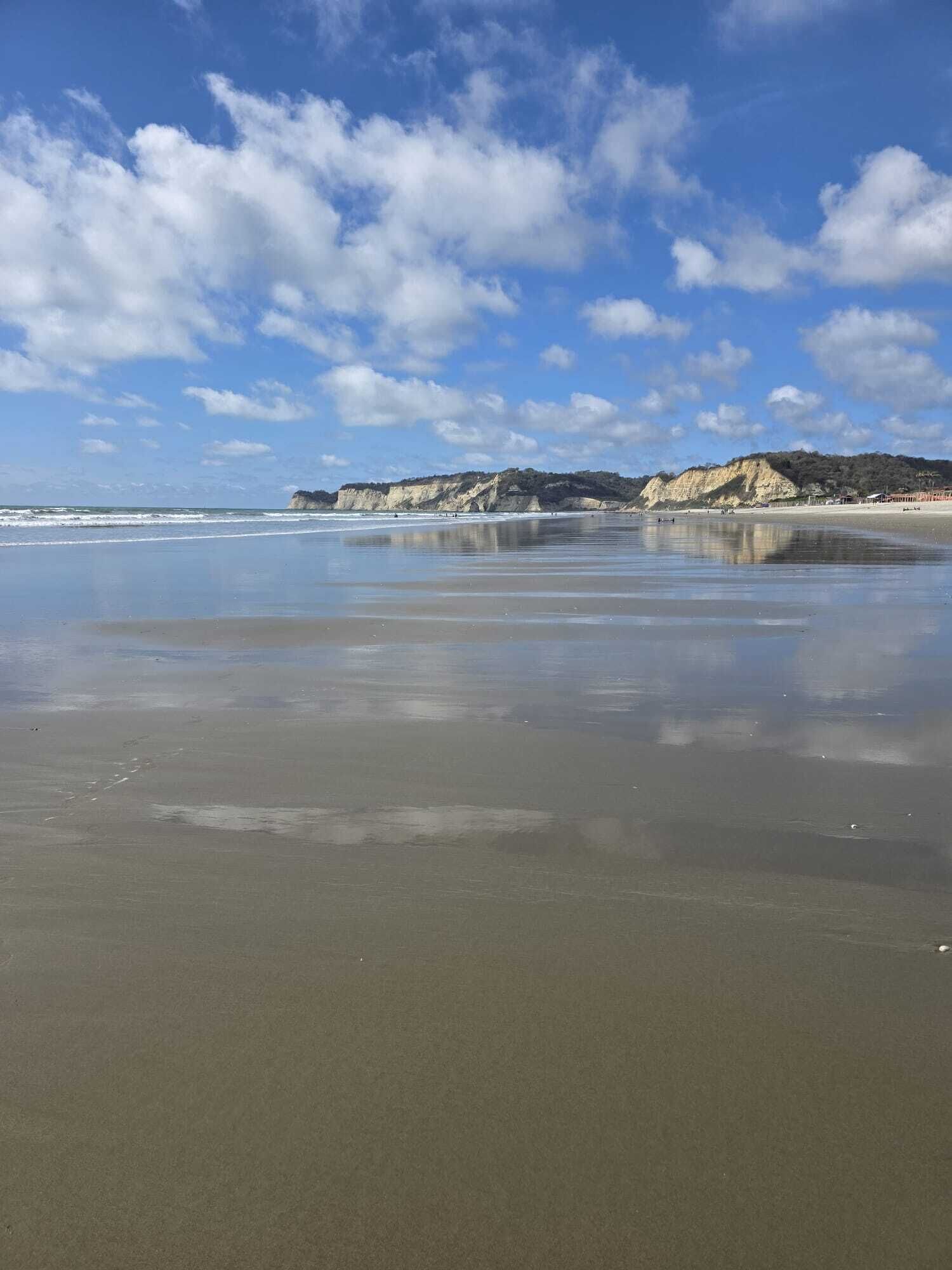 A beach with a clear sky, calm water, and rocky cliffs in the background.