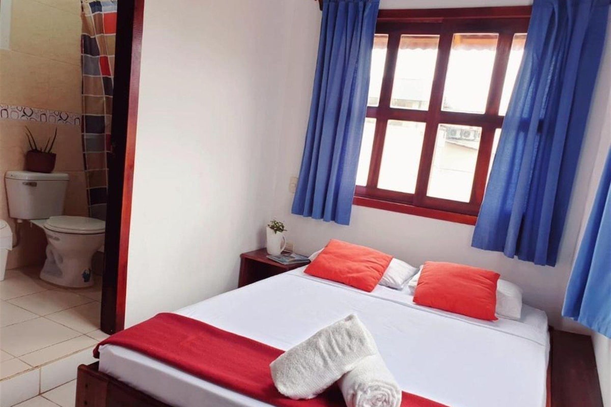 A hotel room with a bed, a window with curtains, a bedside table, and a toilet.