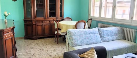 A room with a stone floor, a wooden cabinet, a sofa, a table, and a window with a view of buildings.
