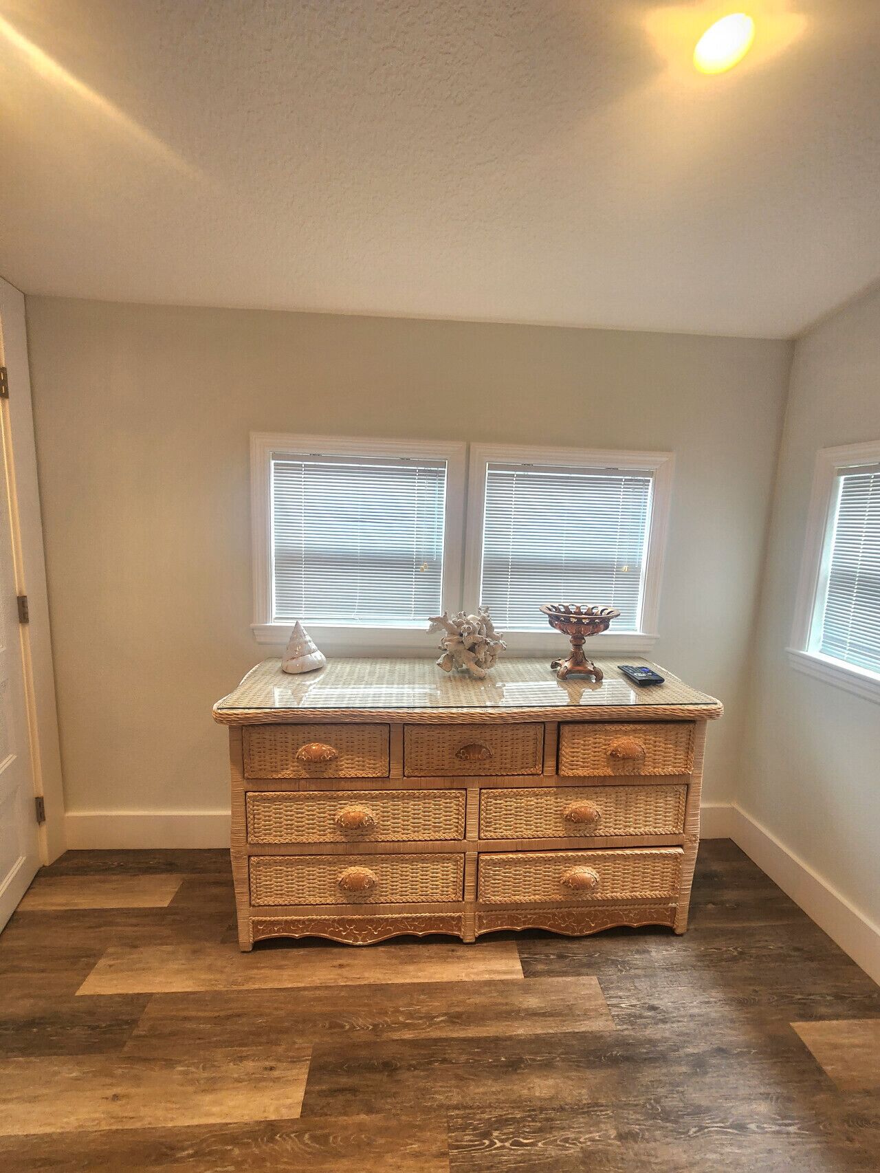 A corner room with a wooden dresser, two windows with blinds, and a light fixture on the ceiling.