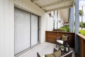 Ein überdachter Balkon mit Schiebeglastür, Korbmöbeln, einem kleinen Tisch mit Kerze und einem Holzpflanzgefäß mit Grünpflanzen.