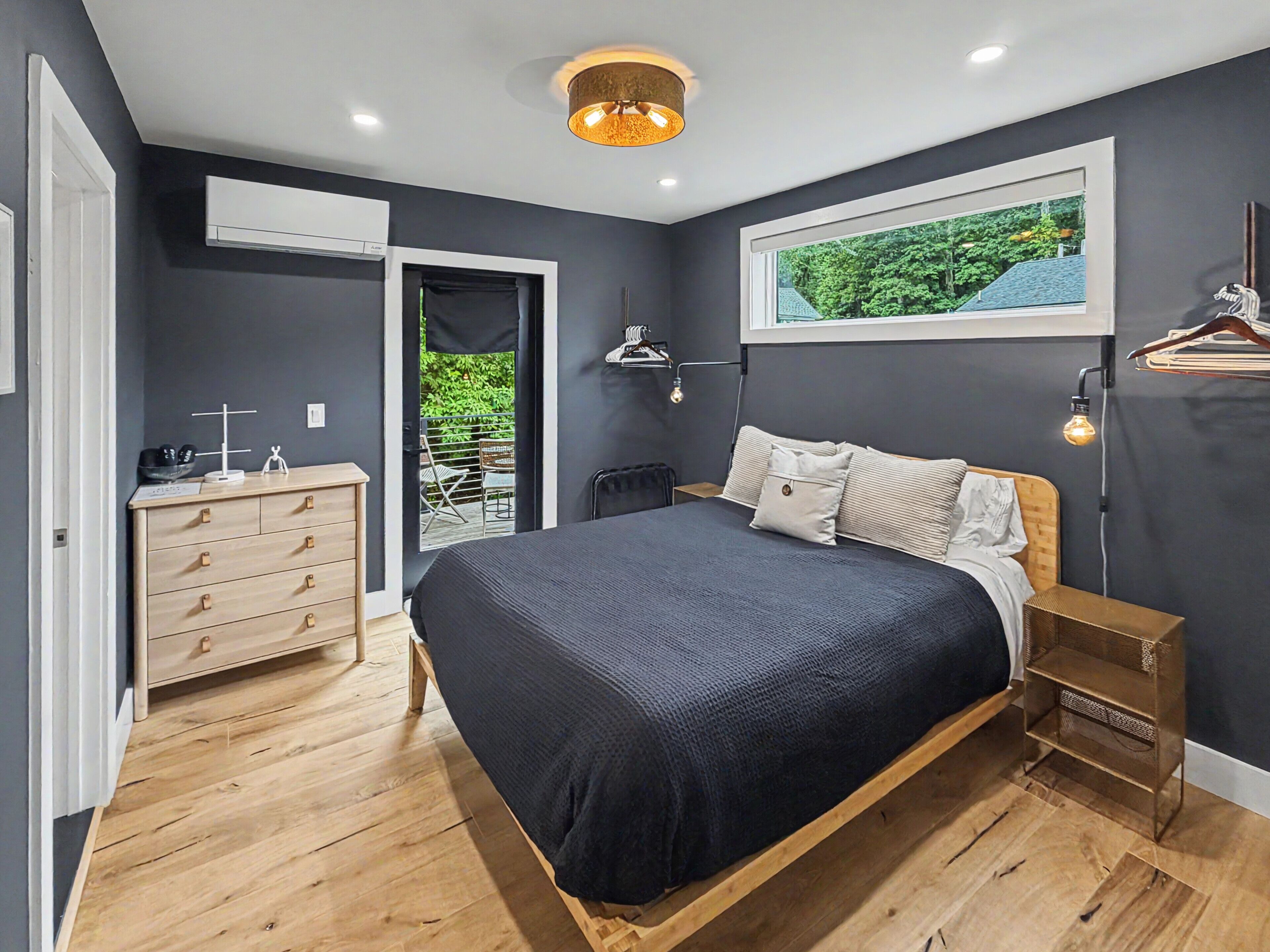 A modern bedroom with a bed, dresser, and a view of greenery through a window.