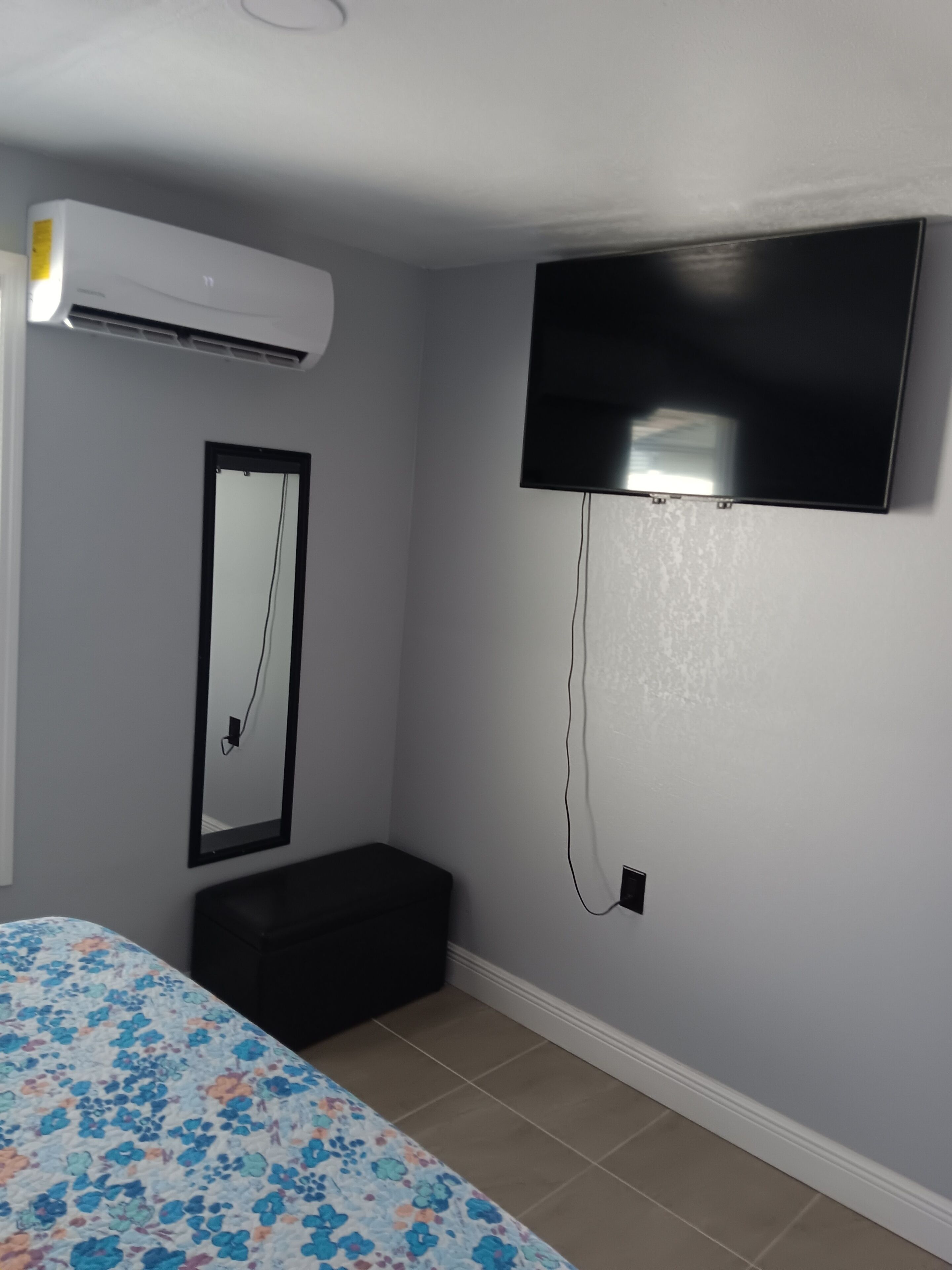 A bedroom with a bed, an air conditioner, a television mounted on the wall, and a small bench.