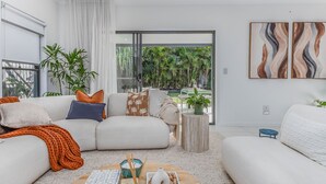 Una sala de estar moderna con un sofá blanco, una mesa de centro con objetos decorativos y vistas a un jardín tropical a través de una puerta de cristal.