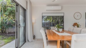 A dining area with a wooden table, white chairs, a large window with a view of greenery, and an air conditioning unit. - Sunrays Sanctuary – 350m to Beach & Pet-Friendly (Casuarina)