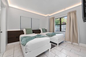 Un dormitorio con dos camas, una ventana con cortinas, techo blanco y televisor montado en la pared.