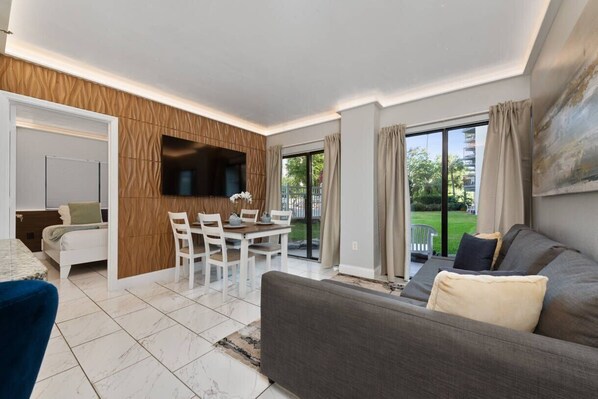 Una sala de estar moderna con un sofá, un televisor montado en la pared, un comedor con sillas y una mesa, y un ventanal con cortinas.