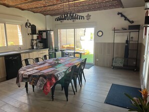Uma área de refeições com uma mesa coberta por um mantelão de retalhos, uma cozinha com geladeira e vista para o exterior.
