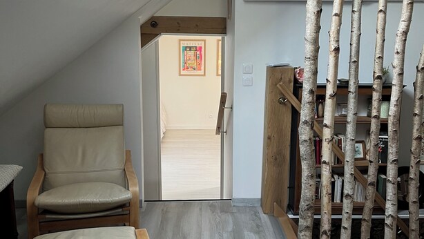 Une pièce dotée d’une lucarne, d’une chaise, d’un banc, d’une bibliothèque en bois et d’une porte donnant sur une autre pièce.