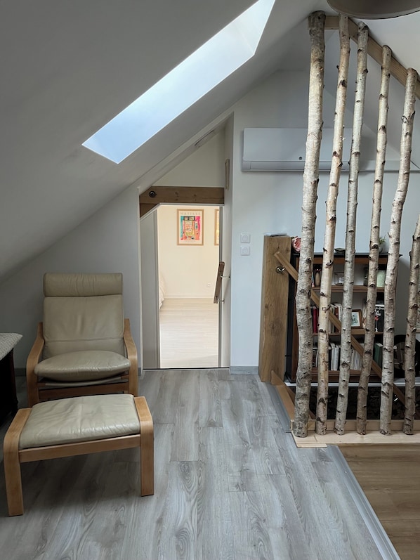 Une piĂšce dotĂ©e dâune lucarne, dâune chaise, dâun banc, dâune bibliothĂšque en bois et dâune porte donnant sur une autre piĂšce.