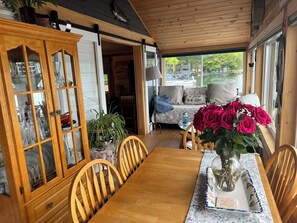 Ett matbord i trä med stolar, ett vitrinskåp med porslin, en soffa med kuddar och en vas med blommor.