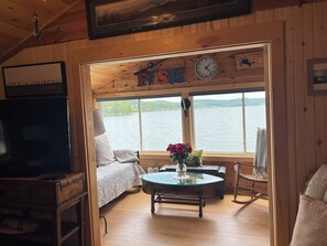 Ein Interieur einer Hütte mit einem Bett, einem Tisch mit einer Vase, einer Uhr und Blick auf einen See.