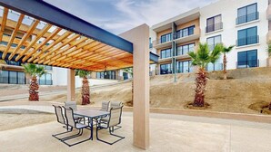 A covered outdoor seating area with a table and chairs, palm trees, and a building with balconies in the background. - Gorgeous Dog-friendly Apartment With Balcony, Open Layout, Heated Pool, & AC (Cabo San Lucas)