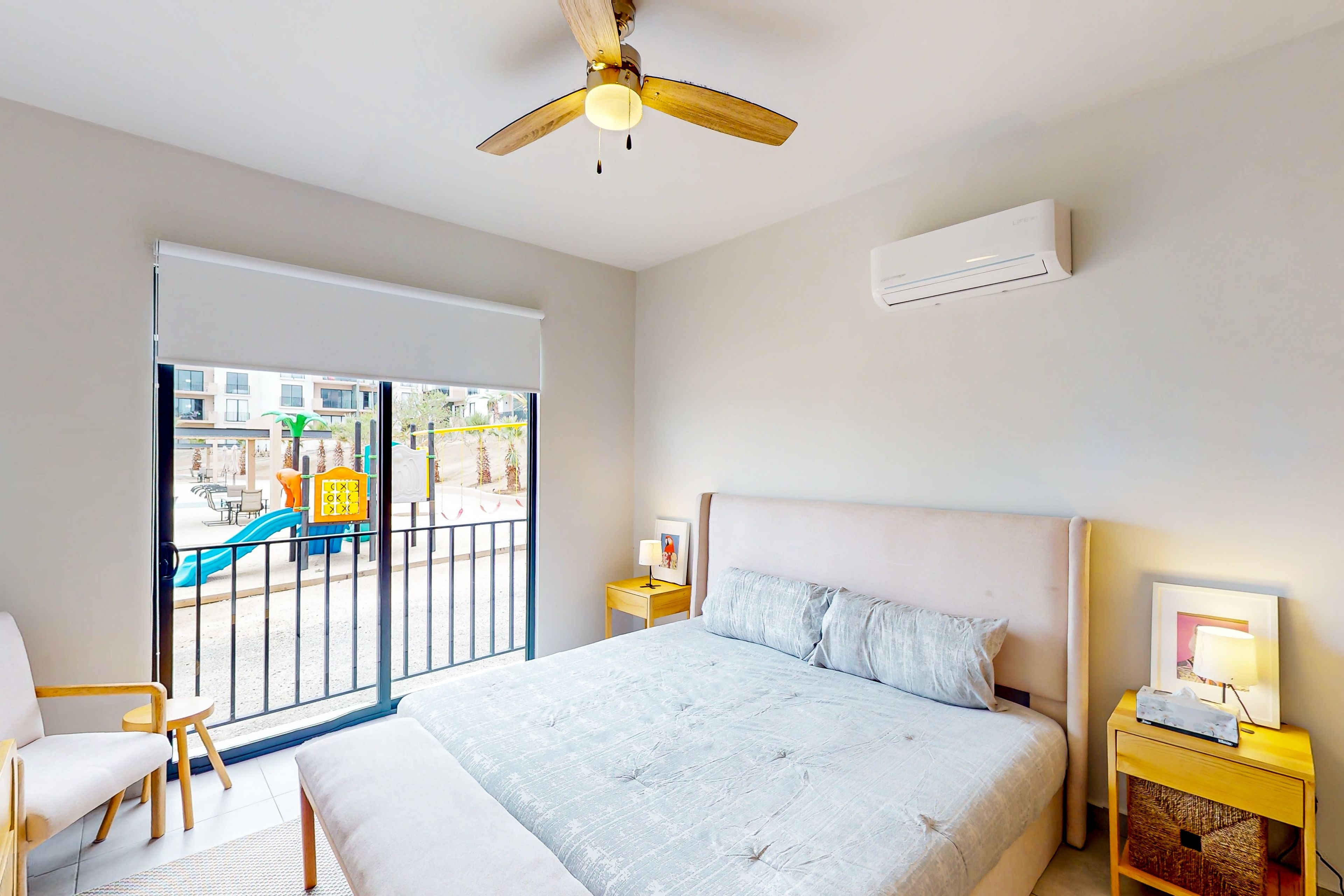A bedroom with a bed, a nightstand, a chair, a ceiling fan, and a view of a playground through a sliding door.