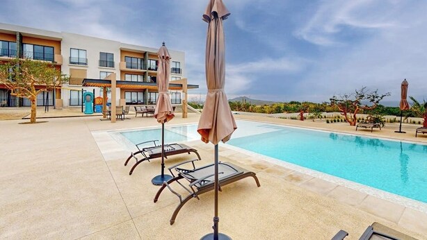 A pool area with lounge chairs and umbrellas, a building with multiple balconies, and a clear sky. - Gorgeous Dog-friendly Apartment With Balcony, Open Layout, Heated Pool, & AC (Cabo San Lucas)