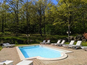 Ein rechteckiger Pool mit einer Pflasterterrasse, umgeben von weißen Liegestühlen und Bäumen im Hintergrund.