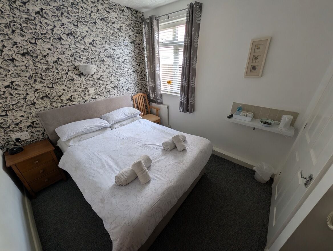 A neatly arranged bedroom with a bed, a nightstand, a chair, a radiator, a window with curtains, and a small shelf with toiletries.