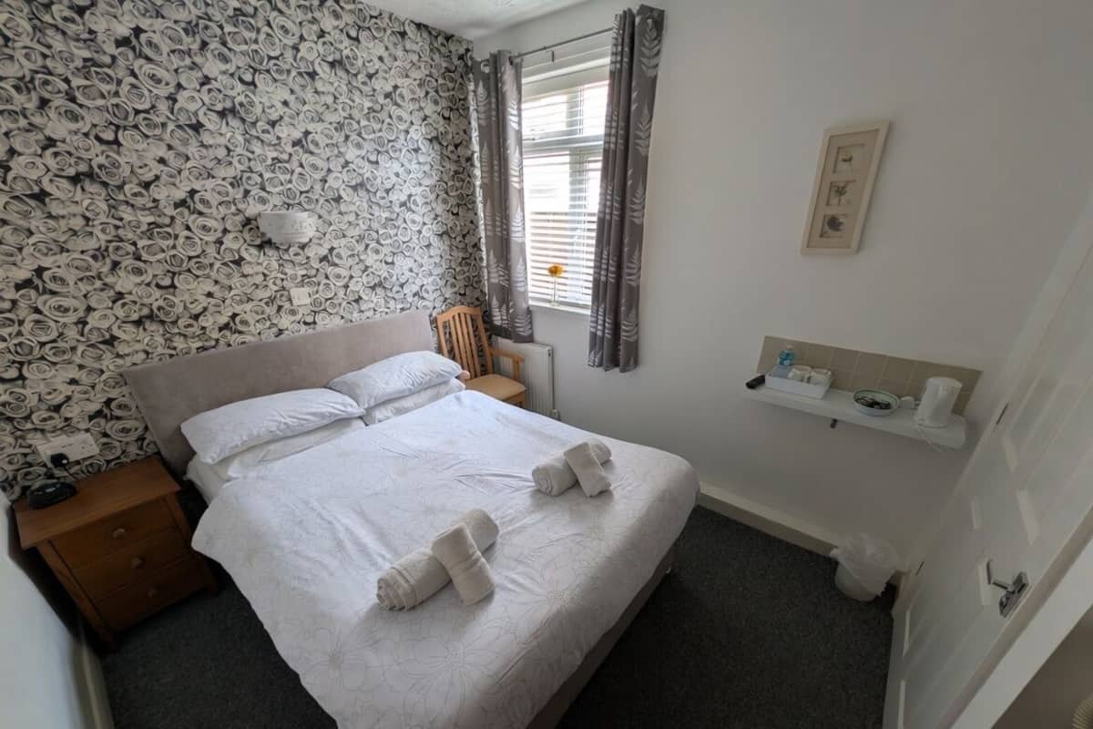 A neatly arranged bedroom with a bed, a nightstand, a chair, a radiator, a window with curtains, and a small shelf with toiletries.