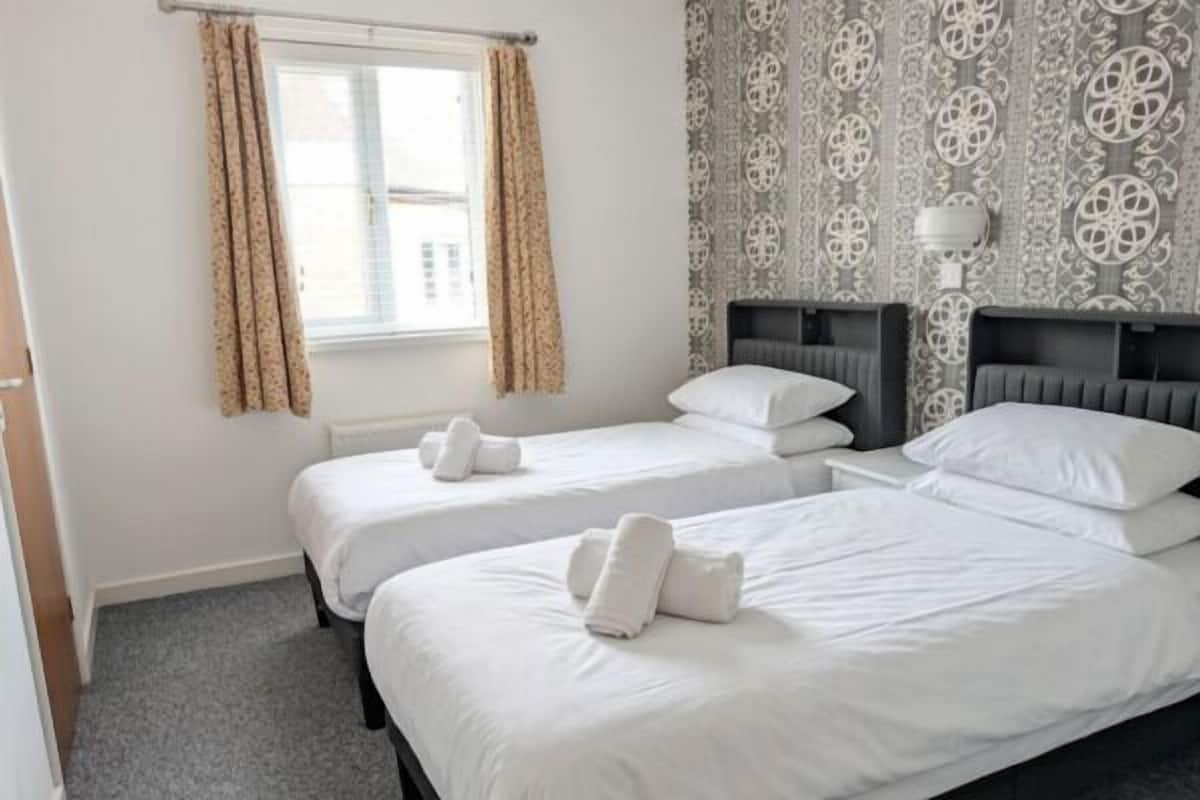 A hotel room with two single beds, a window with curtains, patterned wallpaper, and a bedside lamp.
